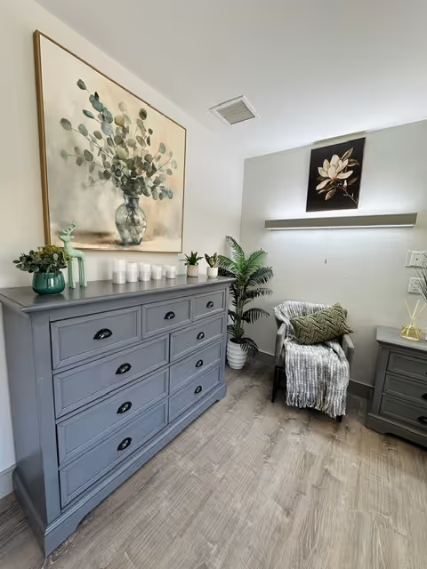 A cozy corner of a room featuring a gray dresser with multiple drawers, decorated with small plants and candles on top. Above the dresser hangs a large framed painting of eucalyptus leaves in a vase. To the right, there is a comfortable armchair draped with a gray knitted throw and a green patterned pillow. A potted plant is placed beside the chair, and a framed floral artwork is mounted on the wall above a narrow shelf. The room has light-colored walls and wood flooring.