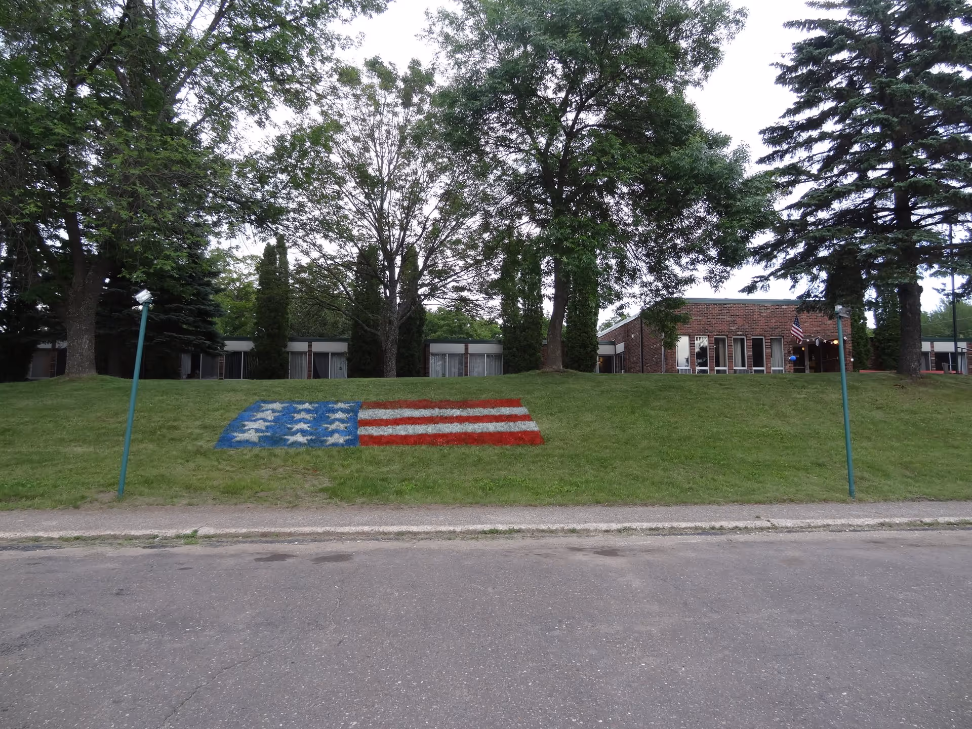 A grassy hill with an American flag painted on it in red, white, and blue. Behind the hill are several trees and a brick building with large windows. Two green poles with lights or lamps are positioned on either side of the hill. The sky is overcast.