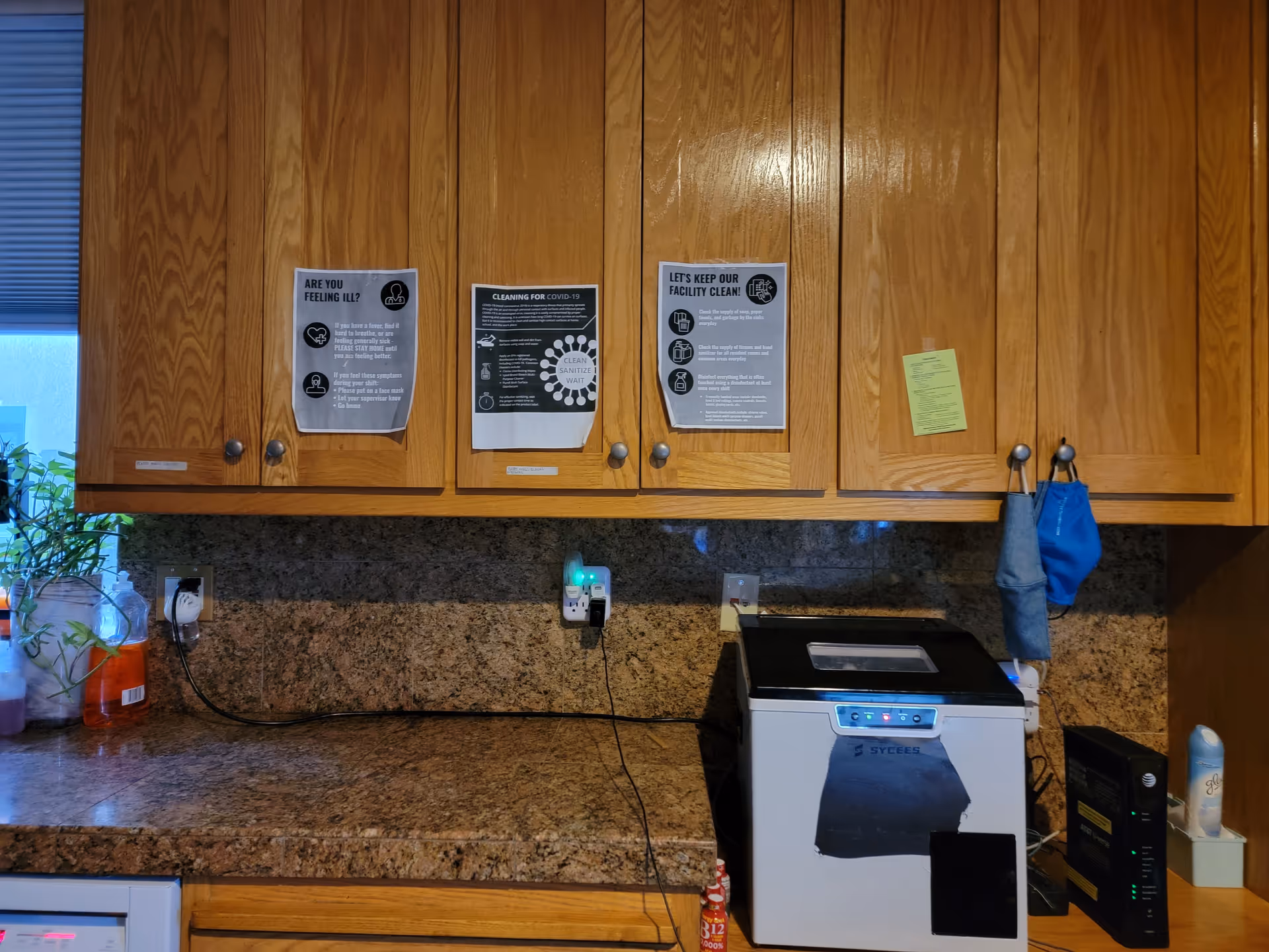 A kitchen countertop with wooden cabinets above it. On the cabinets, there are three printed notices related to health and cleaning protocols. The countertop has a small appliance, a router, a can of air freshener, and some hanging cloths or masks on hooks attached to the cabinet. There is also a plant and some cleaning supplies on the left side near a window with blinds.