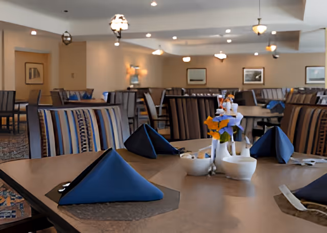 Interior view of a dining room with multiple tables and chairs. Tables are set with blue folded napkins, silverware, and small flower arrangements. The room is softly lit with hanging light fixtures and decorated with framed pictures on the walls.