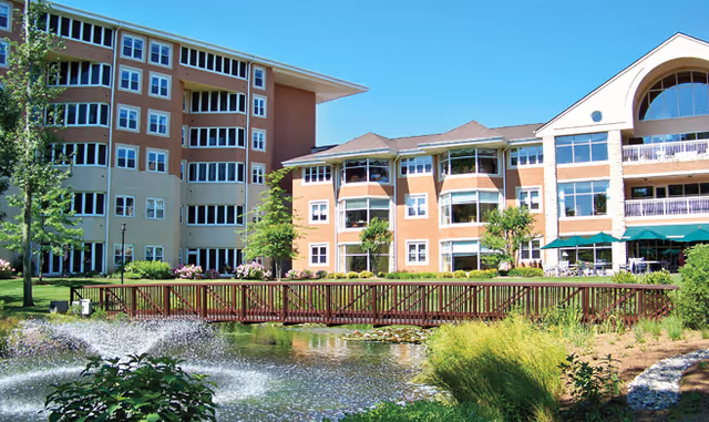 Exterior view of Baywoods of Annapolis senior living facility showing multi-story buildings with balconies, a wooden footbridge over a pond with a water fountain, and landscaped greenery under a clear blue sky.