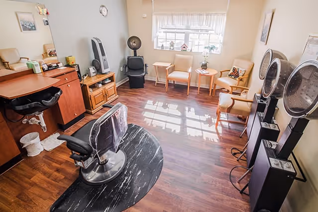 A bright salon room with wooden flooring featuring a black salon chair in front of a wooden counter with a mirror. There are three beige chairs arranged near a window with white curtains, a small round table with a flower vase, and two hair drying machines on the right side of the room.
