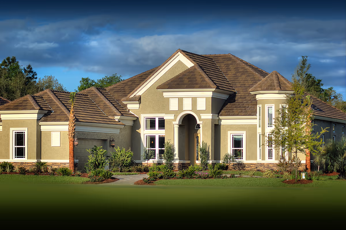 Exterior front view of a large, single-story residential building with a tiled roof, arched entrance, multiple windows, and surrounding landscaping including palm trees and shrubs under a partly cloudy sky.