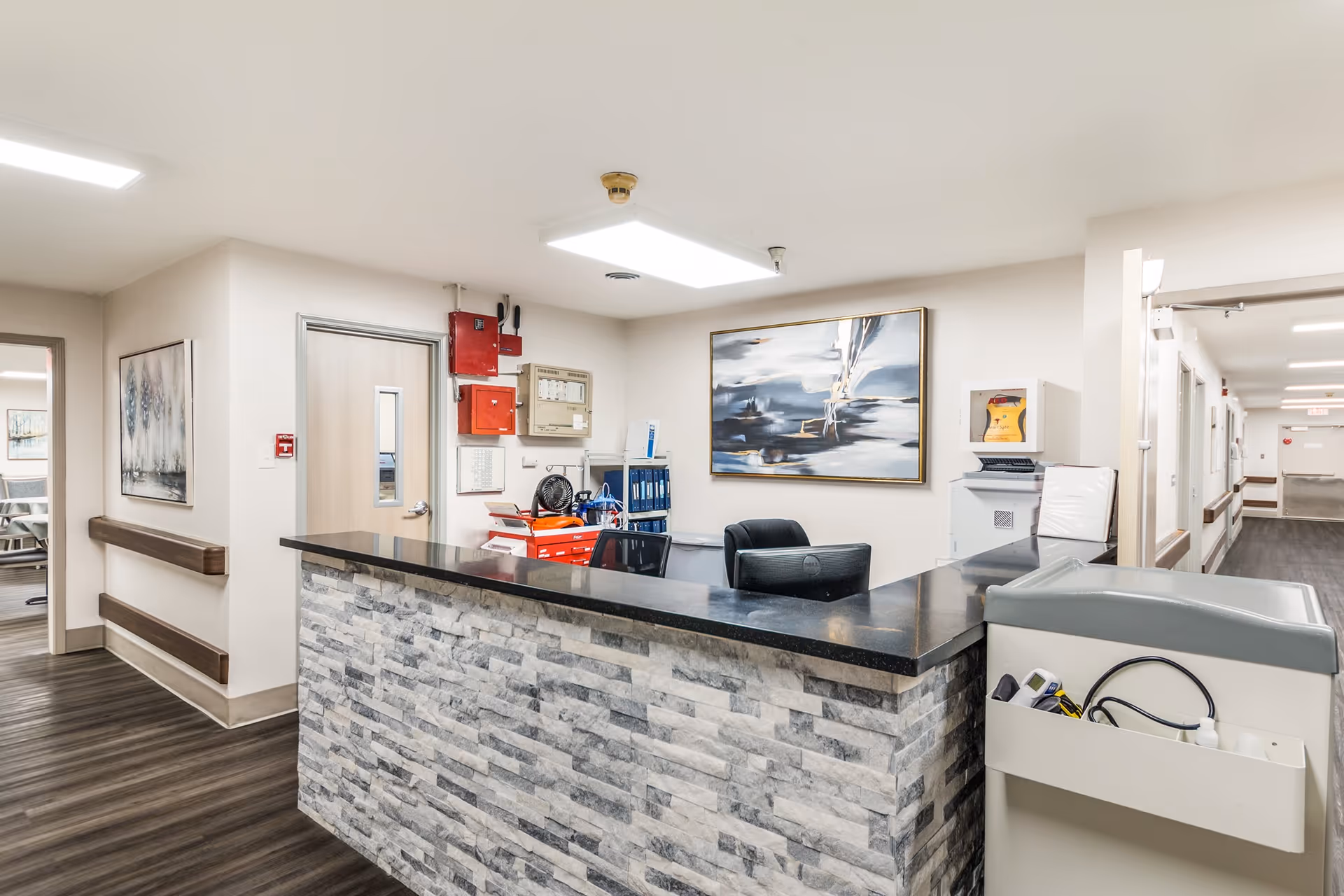 Reception desk area inside a senior care facility with a stone facade counter, black countertop, office chair, and various office supplies. The walls are light-colored with framed artwork, and there is a hallway leading to other rooms.