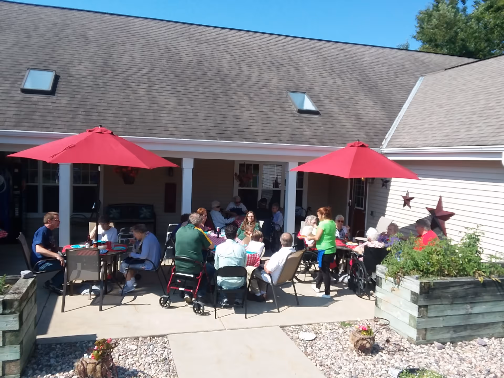 A group of residents and staff seated at patio tables under red umbrellas outside an assisted living facility.