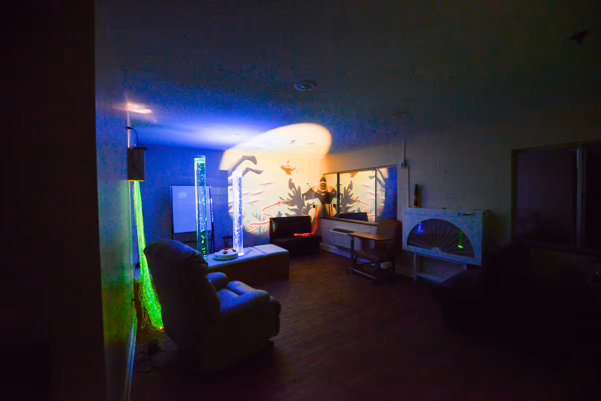 Dimly lit room with a comfortable recliner chair, illuminated bubble tubes, a small table with a chair, and a wall decorated with an underwater-themed mural featuring fish and coral.