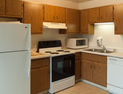 A kitchen with wooden cabinets, a white refrigerator, an electric stove with oven, a microwave, a double sink, and a dishwasher. The countertop is light-colored, and there is a soap dispenser mounted on the wall above the sink.