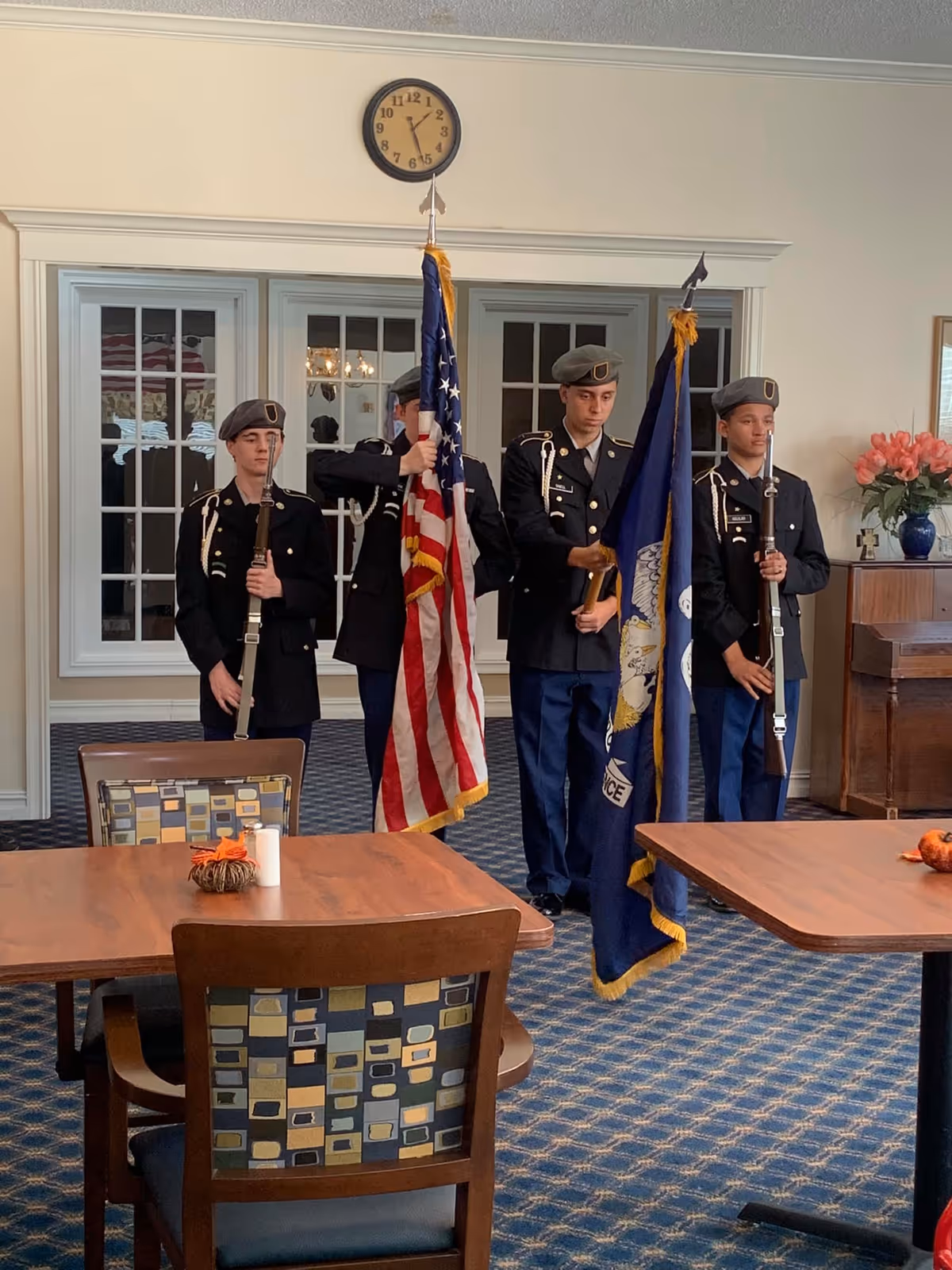 Four young men in military uniforms standing indoors holding flags, including the American flag and a blue flag with an eagle emblem. They are standing in a room with patterned carpet, wooden tables and chairs, a clock on the wall, and a piano with a vase of flowers on top.