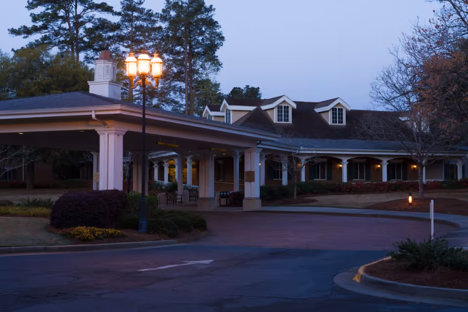 Exterior view of a senior living facility named Brandon Wilde at dusk, showing a covered entrance with columns, a lit street lamp, and a building with dormer windows surrounded by trees and landscaping.