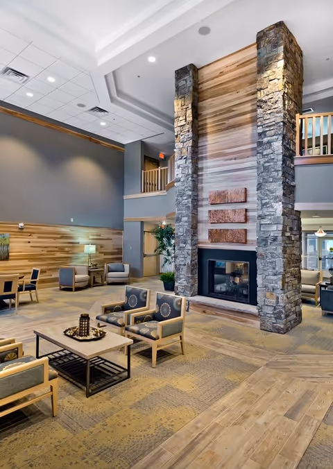 A spacious and well-lit living room area in a senior living facility featuring a tall stone and wood fireplace as the focal point. The room has comfortable chairs arranged around a coffee table, a patterned carpet, wooden wall paneling, and a high ceiling with recessed lighting. There is a second-floor balcony visible above the fireplace and a few potted plants adding greenery to the space.