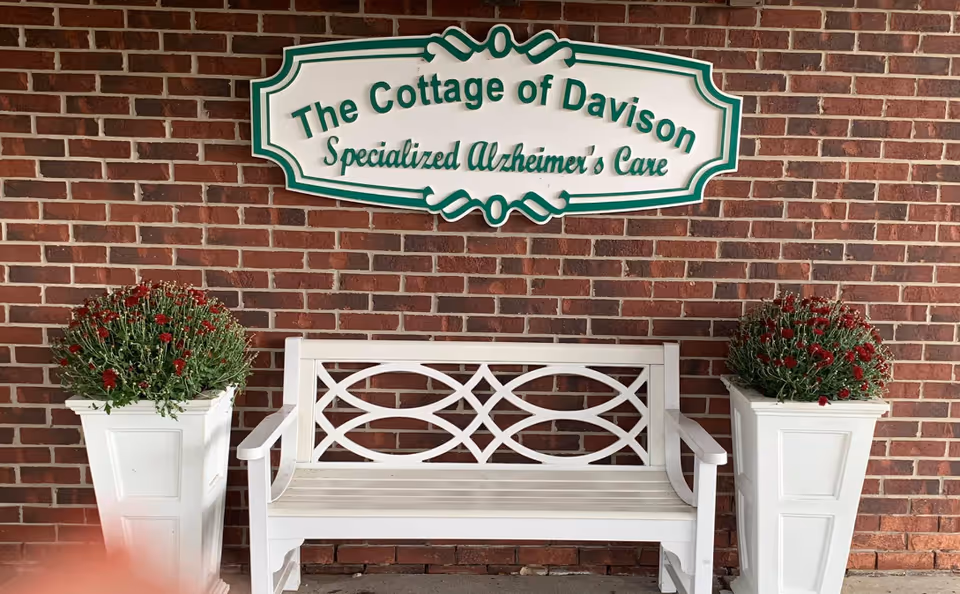 White wooden bench placed against a red brick wall with a sign above it that reads 'The Cottage of Davison Specialized Alzheimer's Care'. On either side of the bench are white planters with red flowers.
