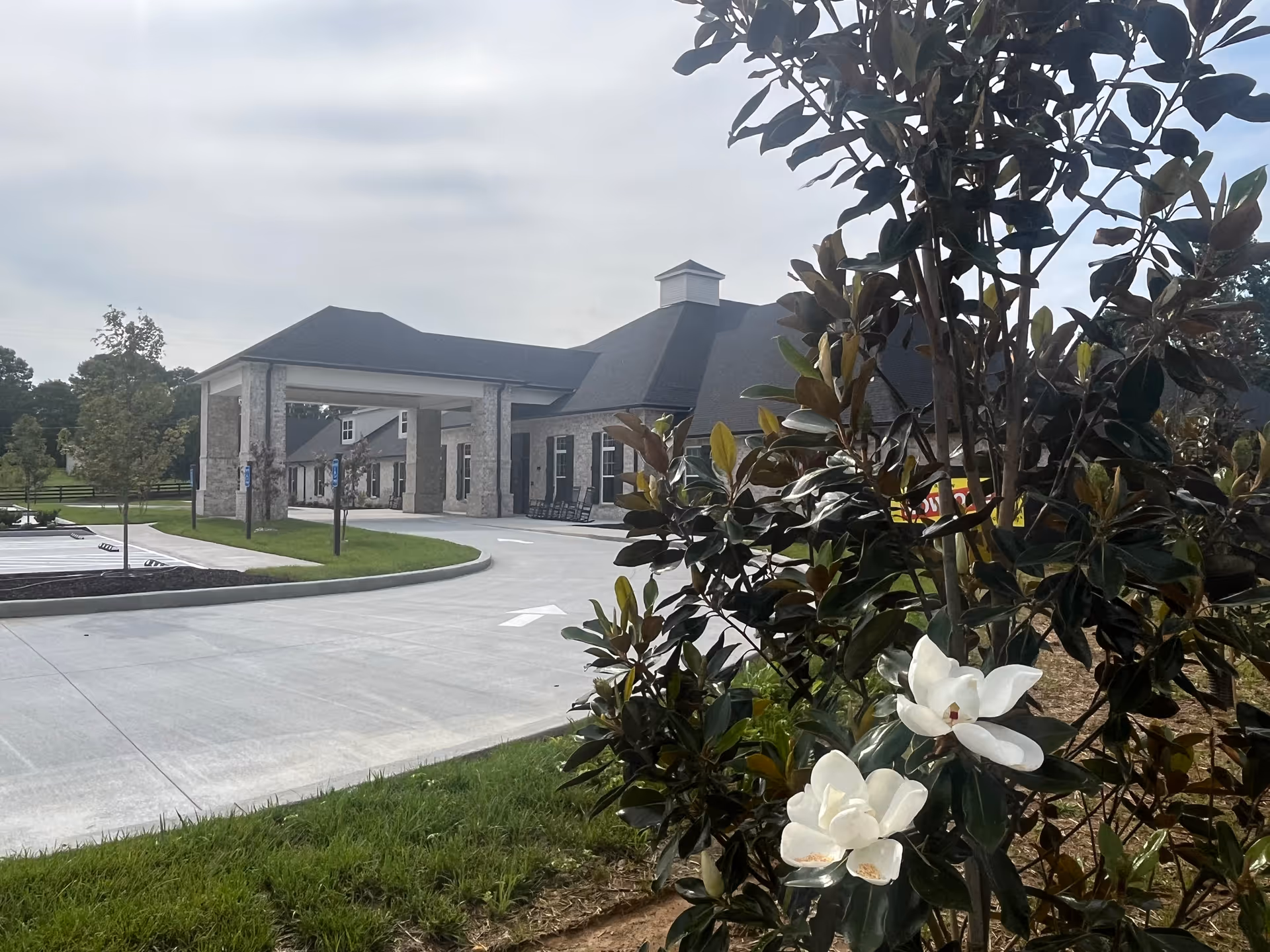 Exterior view of a senior living facility building with a covered entrance and a driveway. In the foreground, there is a flowering tree with white blossoms. The sky is partly cloudy.