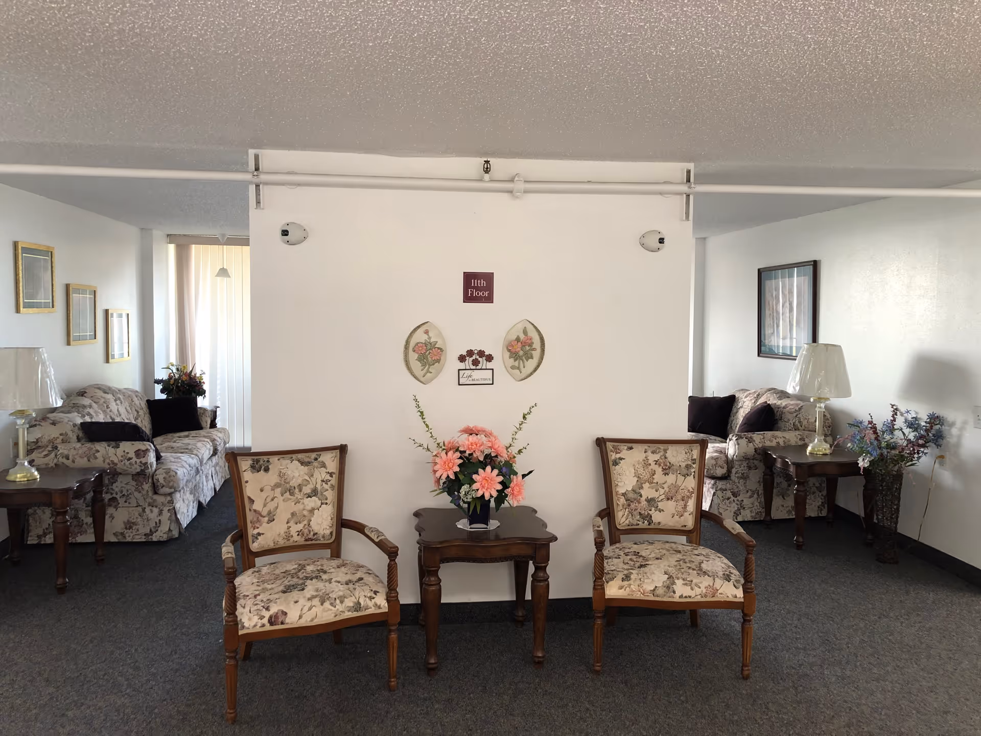A sitting area on the 11th floor of Fellowship Manor featuring two floral upholstered armchairs with wooden frames placed on either side of a small wooden table holding a vase of pink flowers. Behind the chairs is a white wall with floral decorations and a sign that reads '11th Floor'. On both sides of the wall, there are floral patterned sofas, side tables with lamps, framed pictures on the walls, and additional flower arrangements.