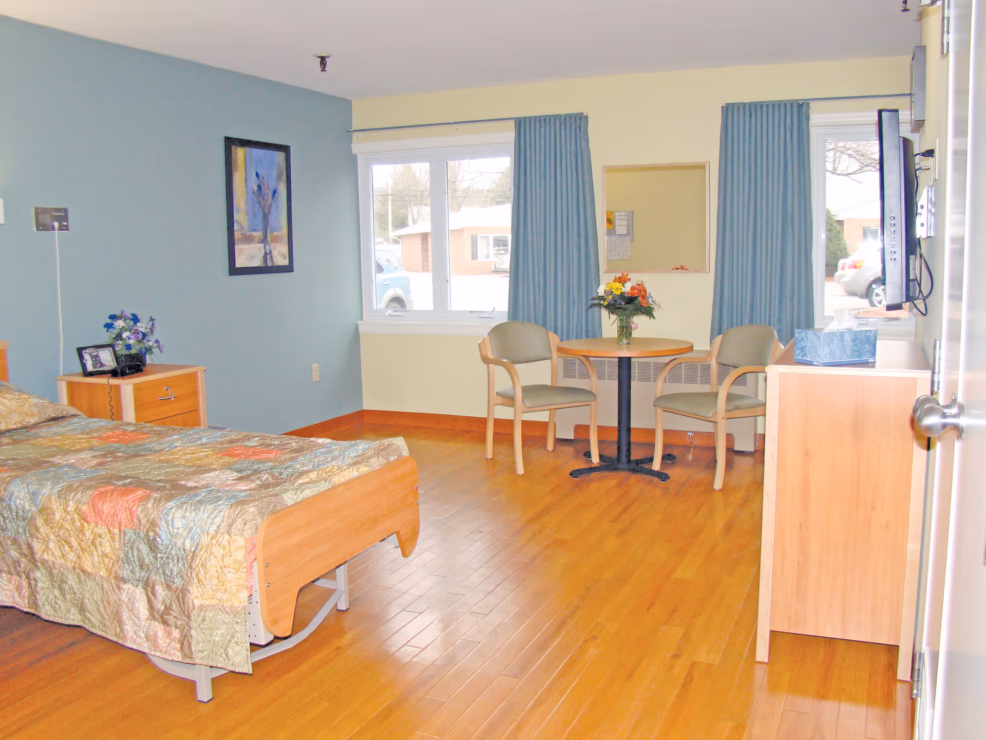 A bright and clean bedroom in a senior living facility with a single bed covered by a colorful quilt, a wooden nightstand with a small flower arrangement and a framed photo, two chairs and a round table with a flower vase near two windows with blue curtains, and a wall-mounted TV on the right side.