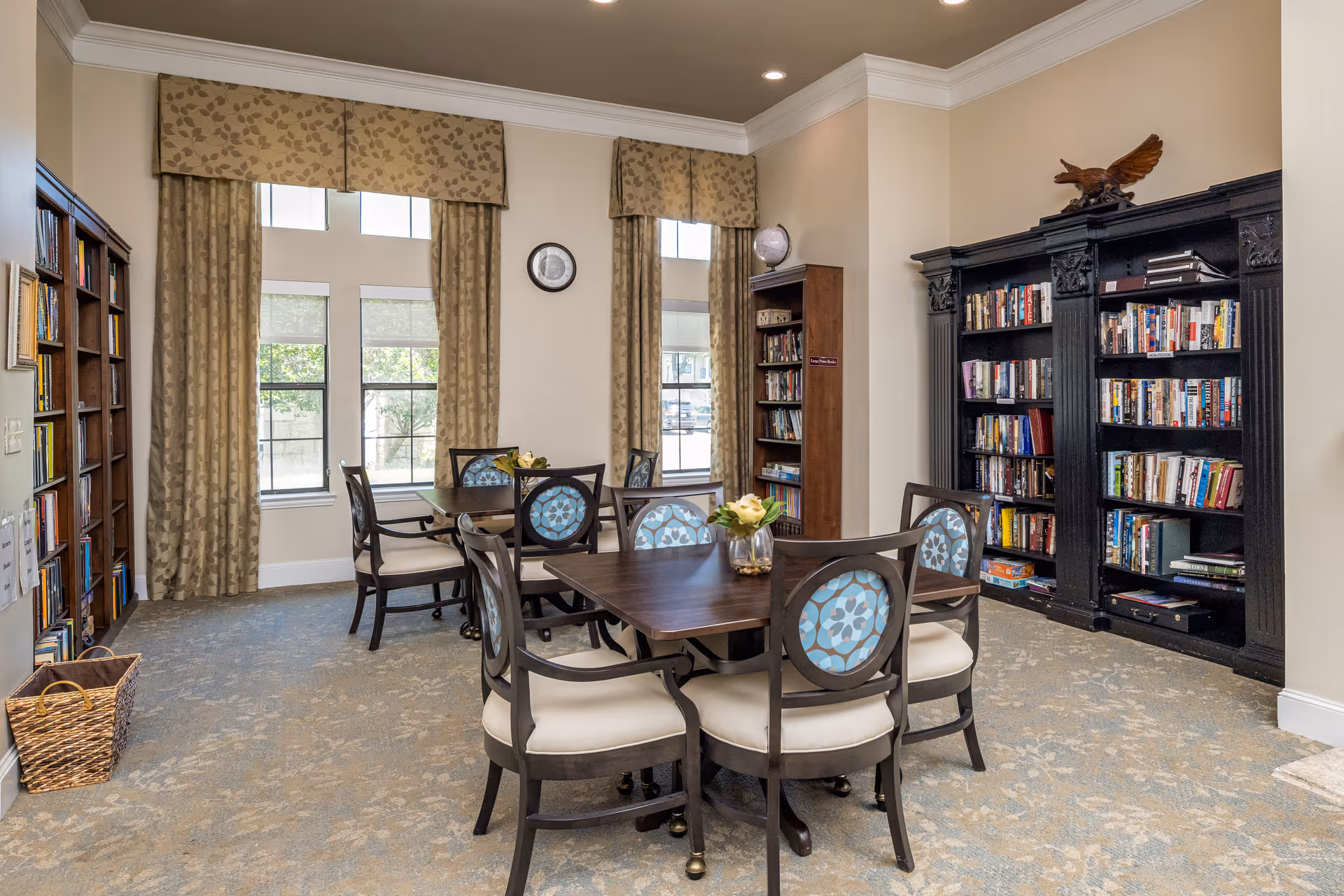A cozy reading and activity room with two wooden tables surrounded by chairs with patterned blue backs and cream seats. The room has large windows with beige patterned curtains, a clock on the wall, and several bookshelves filled with books. A decorative eagle sculpture is placed on top of a large black bookshelf.