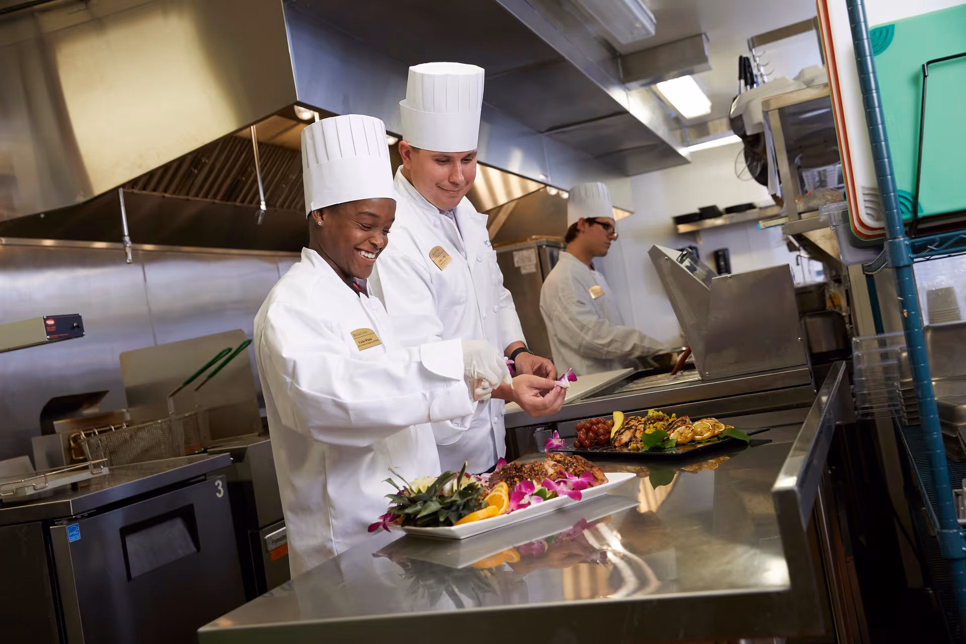 Three chefs in white uniforms and tall hats preparing and plating garnished dishes in a commercial kitchen.