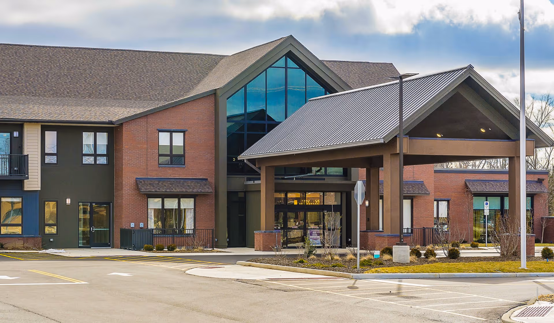 Exterior view of The Patterson senior living facility showing a modern two-story building with a large covered entrance, brick and dark green walls, multiple windows, and a parking area in front under a cloudy sky.