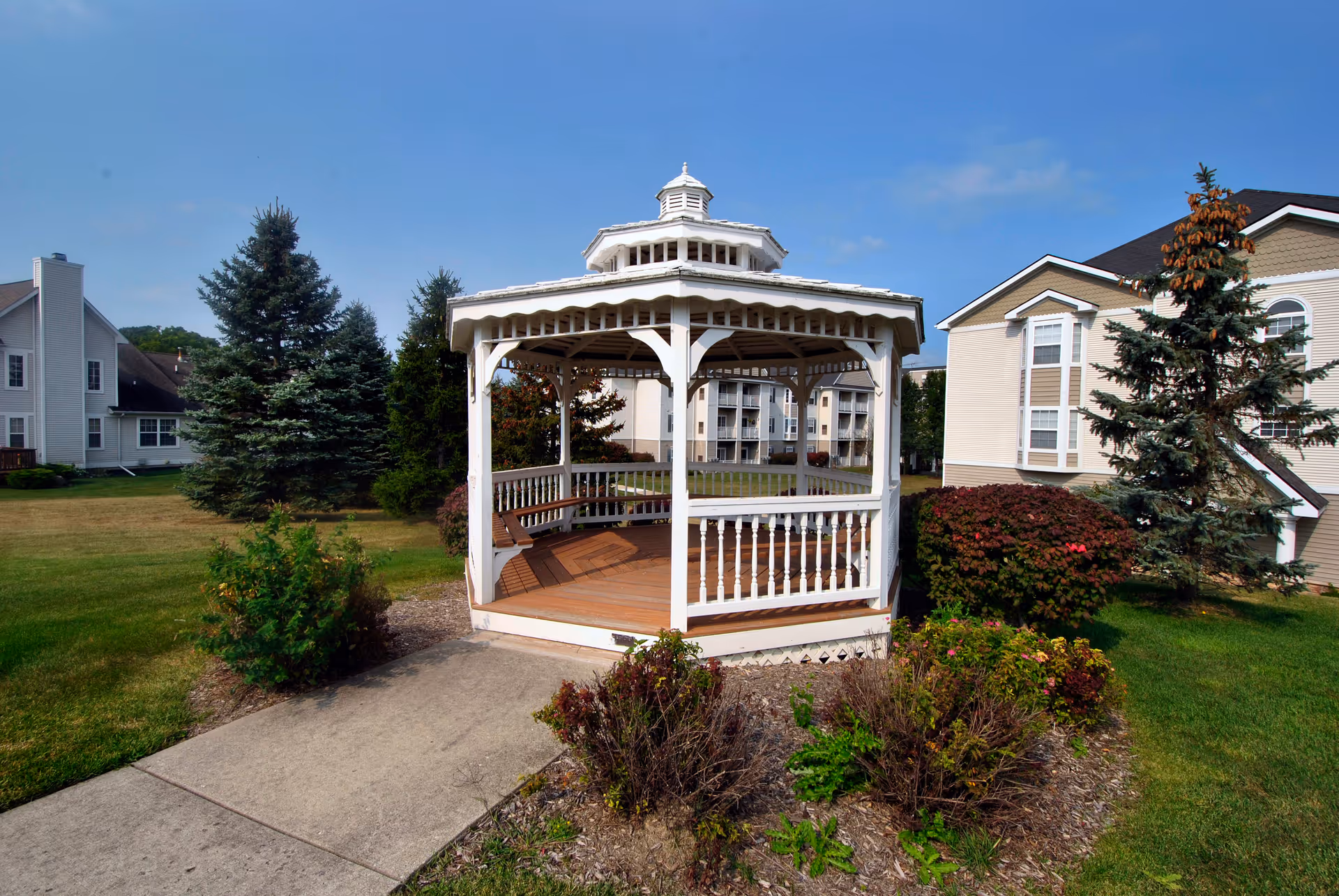 A white wooden gazebo with a shingled roof situated on a concrete path surrounded by green grass, bushes, and trees, with residential buildings in the background under a clear blue sky.
