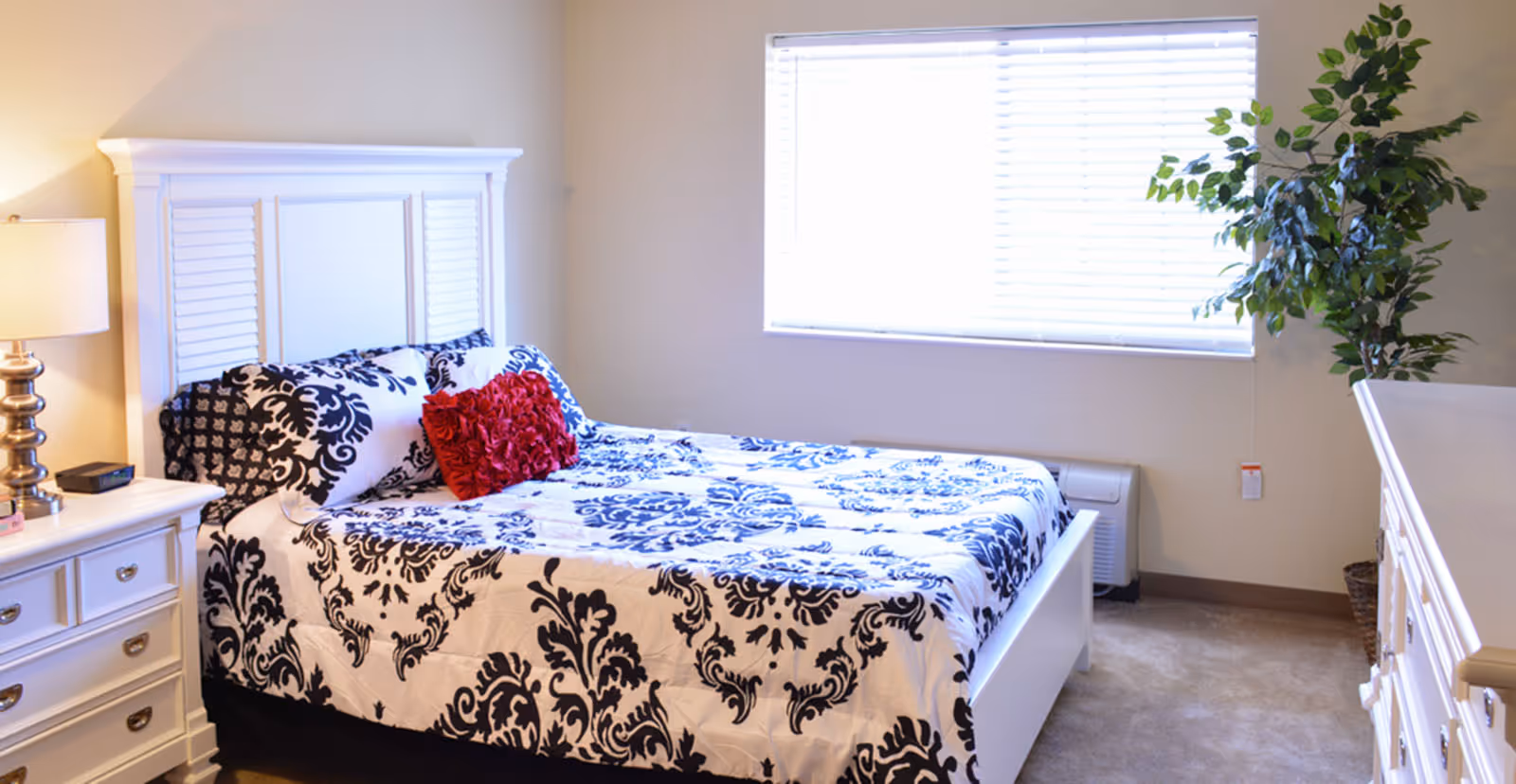 Bright bedroom with a white headboard, black-and-white patterned bedding with a red accent pillow, nightstand, dresser, and a window with blinds.