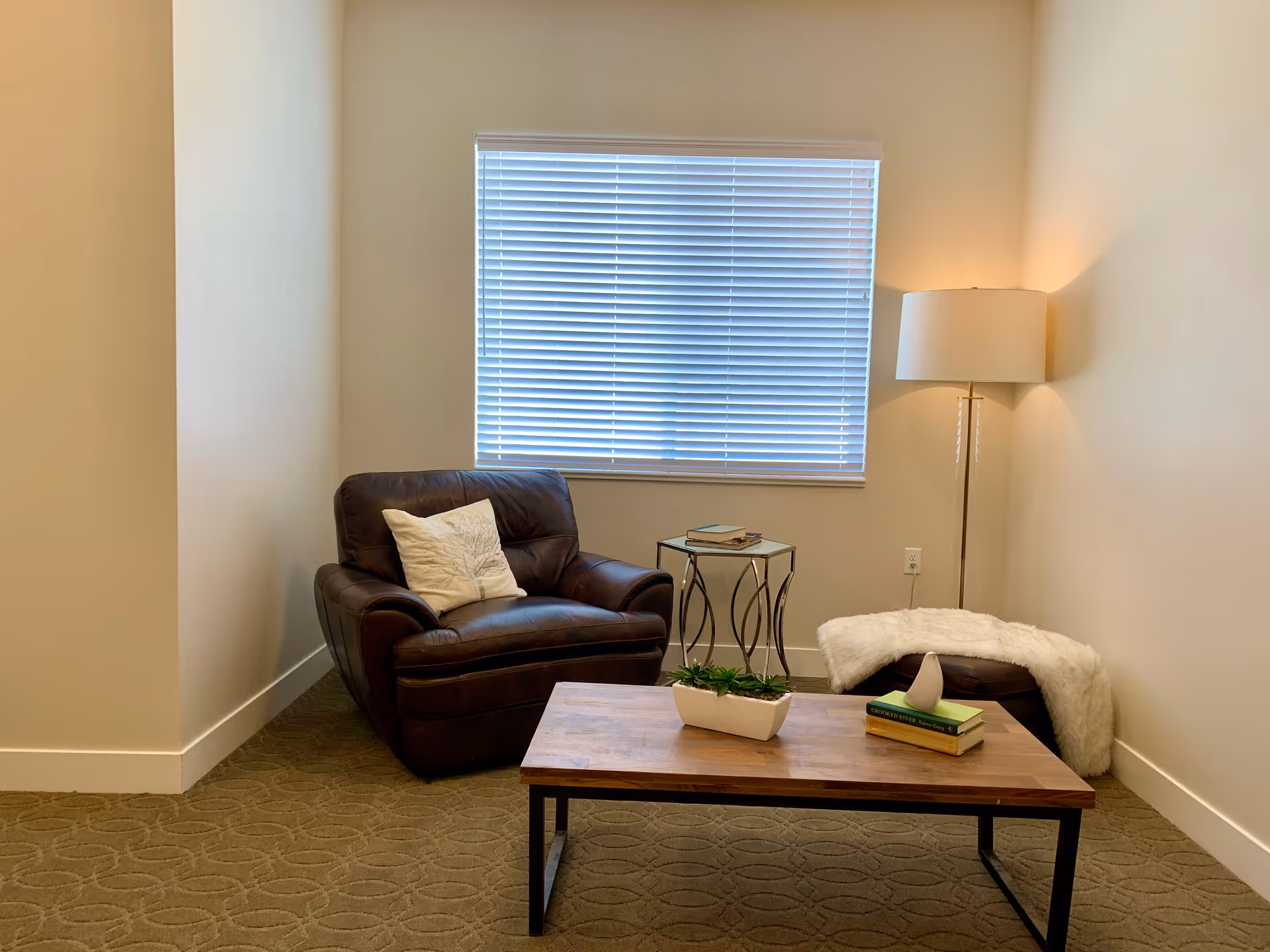 A cozy sitting area with a dark brown leather armchair with a white pillow, a wooden coffee table with a small plant and books, a small side table with books, a floor lamp, and a window with closed blinds in a room with beige walls and carpeted floor.