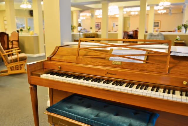 Upright wooden piano with a padded bench in the foreground of a bright communal lounge with chairs and tables in the background.