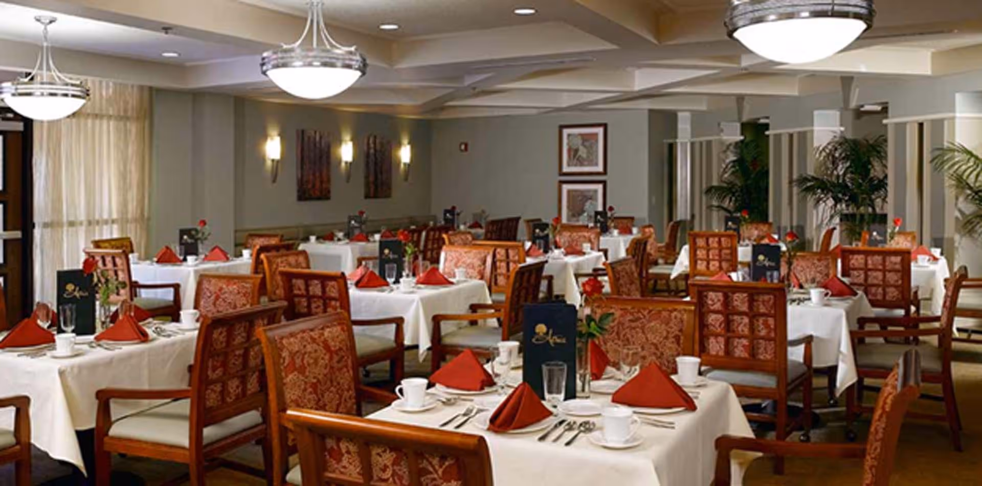 Spacious dining room with multiple tables set with white tablecloths, red napkins, and wooden chairs.