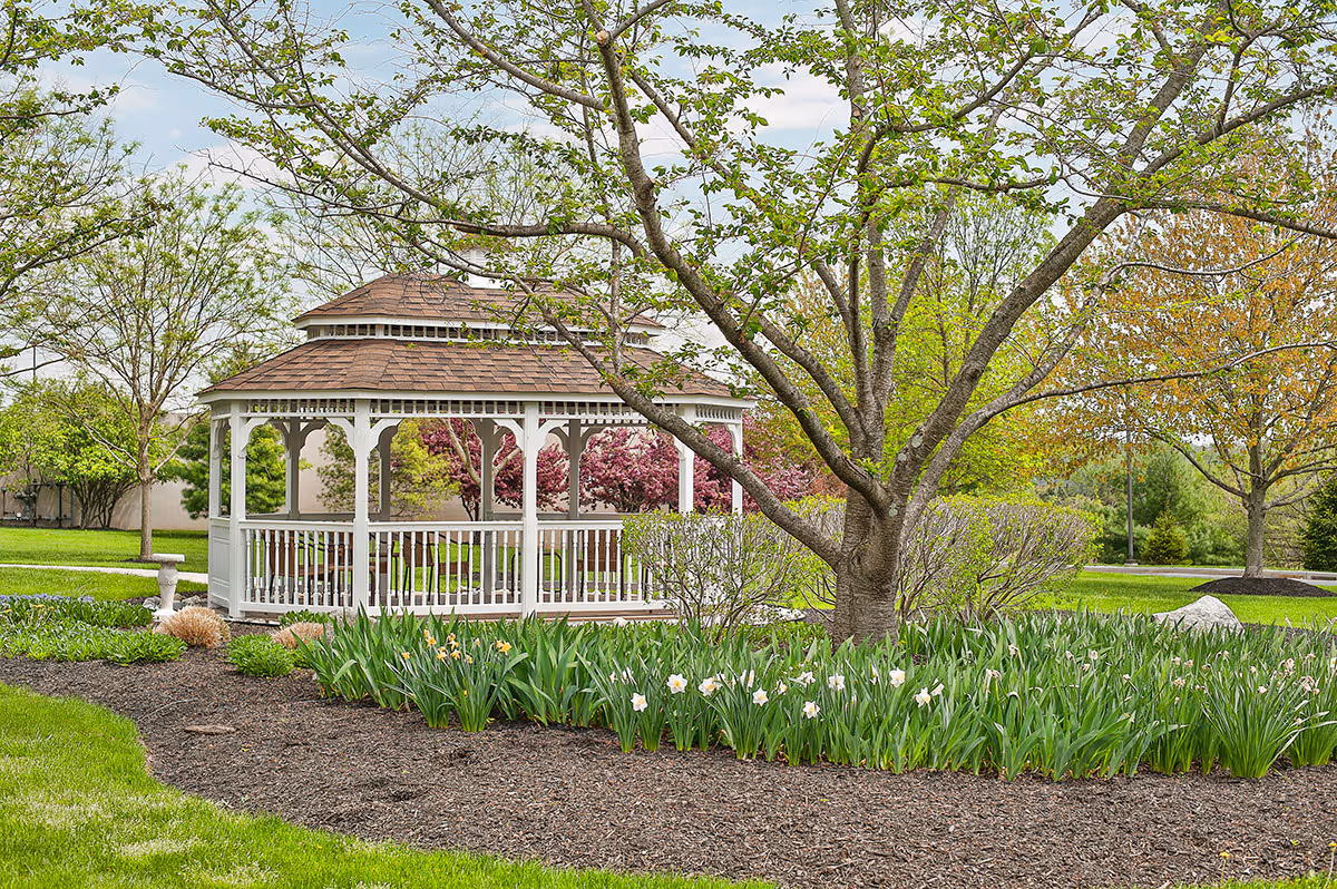 A white wooden gazebo amid trees, flowering beds, and green lawn in a landscaped outdoor area.