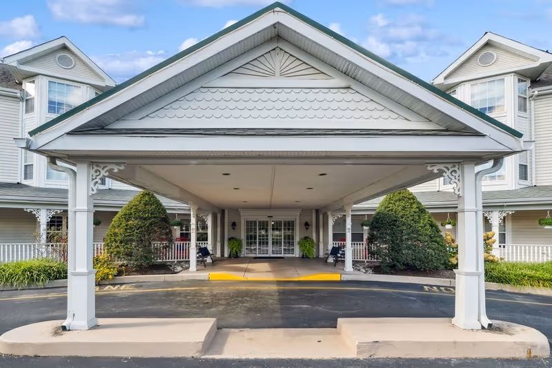 Entrance of a senior living facility with a covered drop-off area supported by white pillars, landscaped bushes on either side, and glass double doors leading inside.