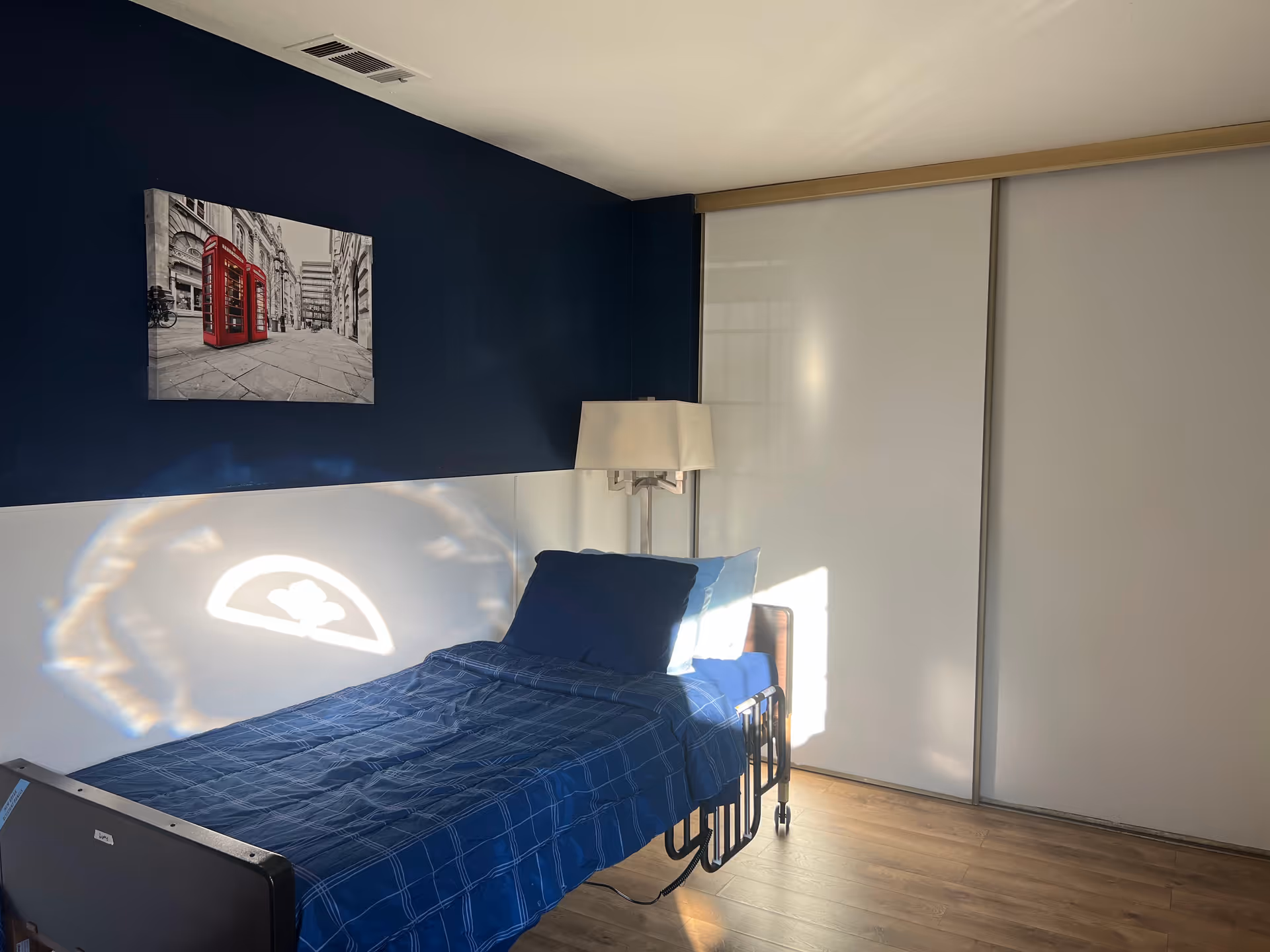 A bedroom with a single bed covered in blue bedding, a bedside lamp, and a wall painting of red telephone booths in a black and white cityscape. The room has a dark blue accent wall and white sliding closet doors.