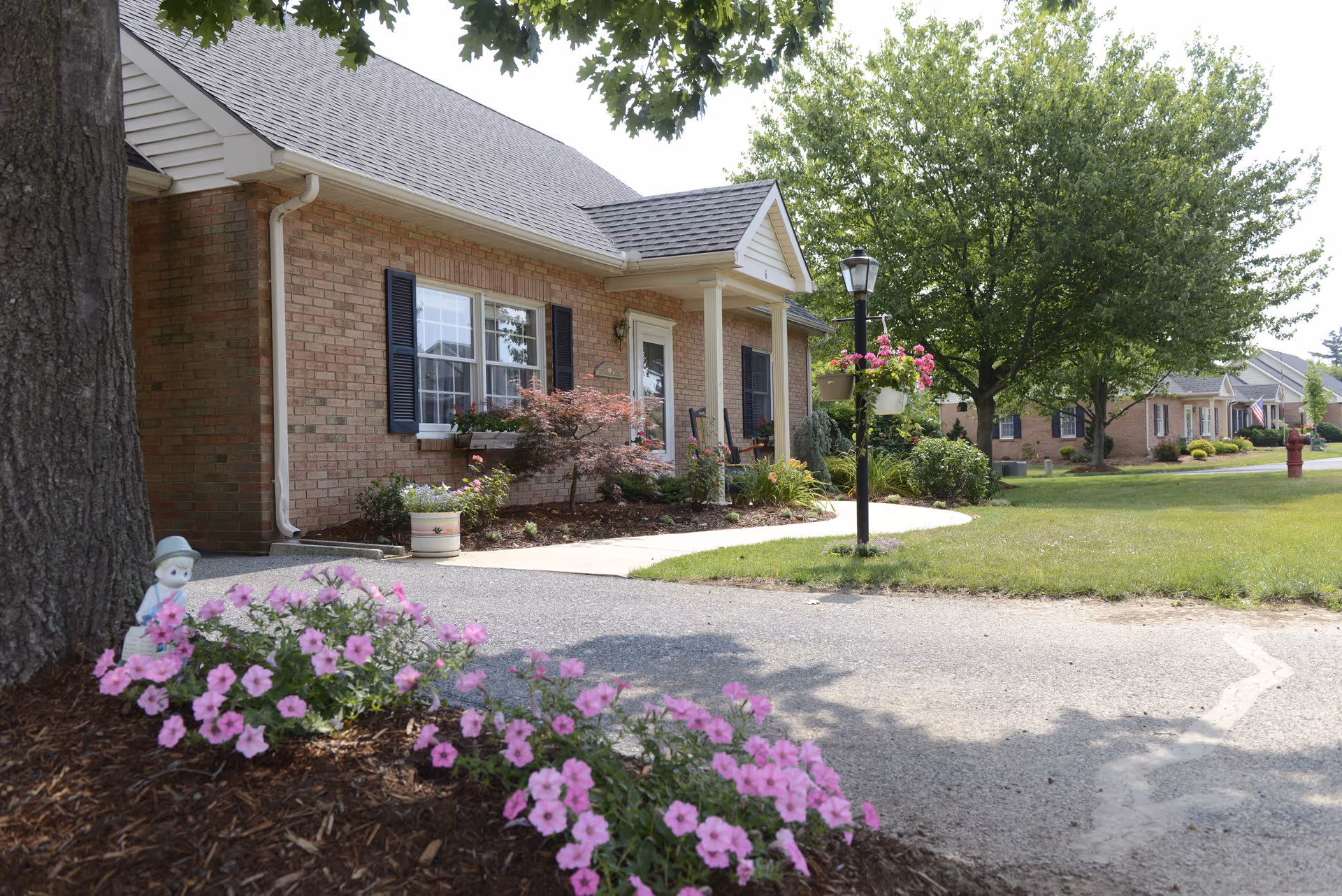 Exterior view of a single-story brick residential building with a small porch, black shutters, and a white door. The front yard features a garden with pink flowers, a tree, and a lamp post with hanging flower baskets. Other similar buildings and green lawns are visible in the background under a clear sky.