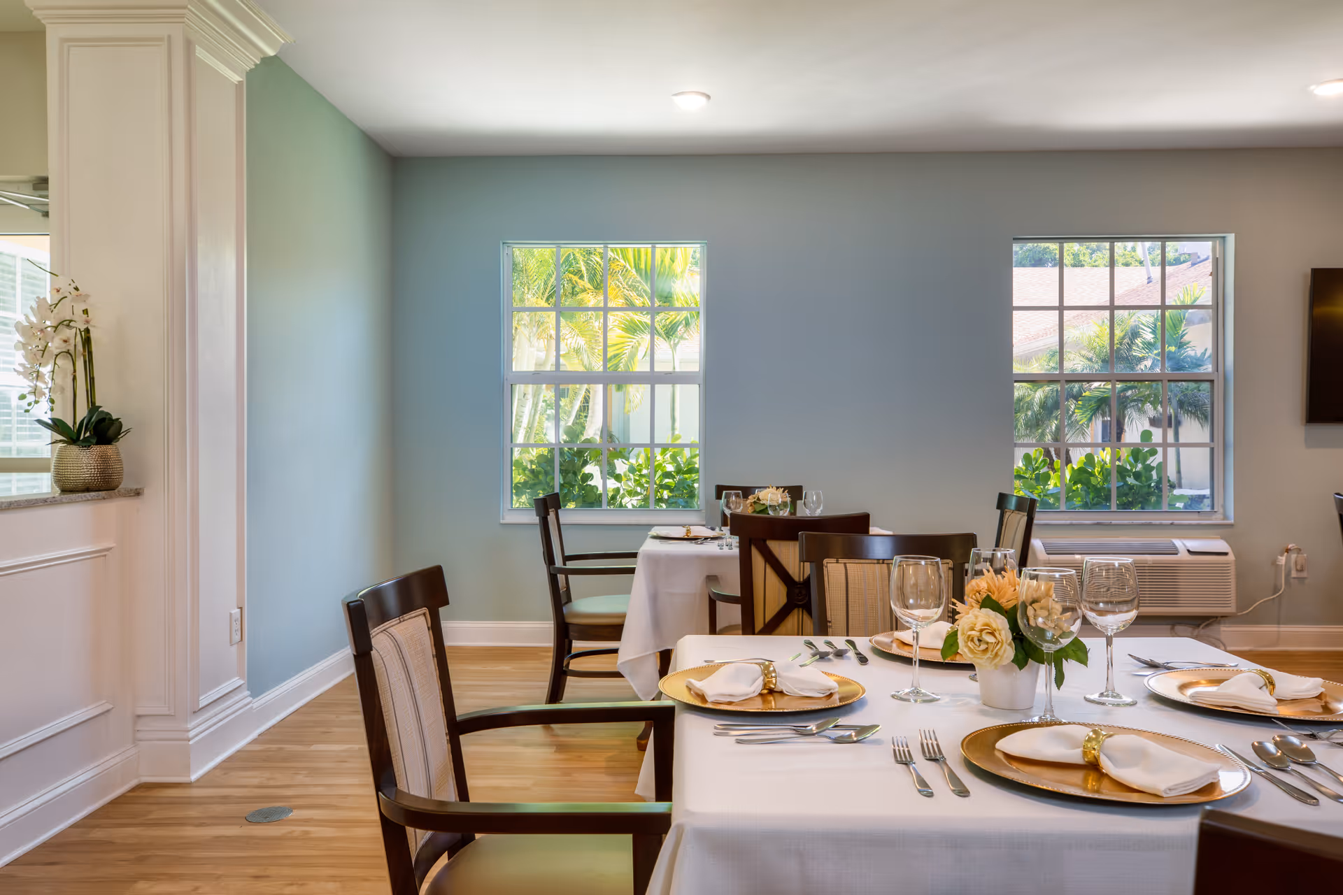 A bright dining room with wooden floors and light blue walls featuring two windows with views of greenery outside. The room has tables set with white tablecloths, gold-rimmed plates, neatly folded napkins with napkin rings, wine glasses, and a small floral centerpiece. Wooden chairs with cushioned seats surround the tables.