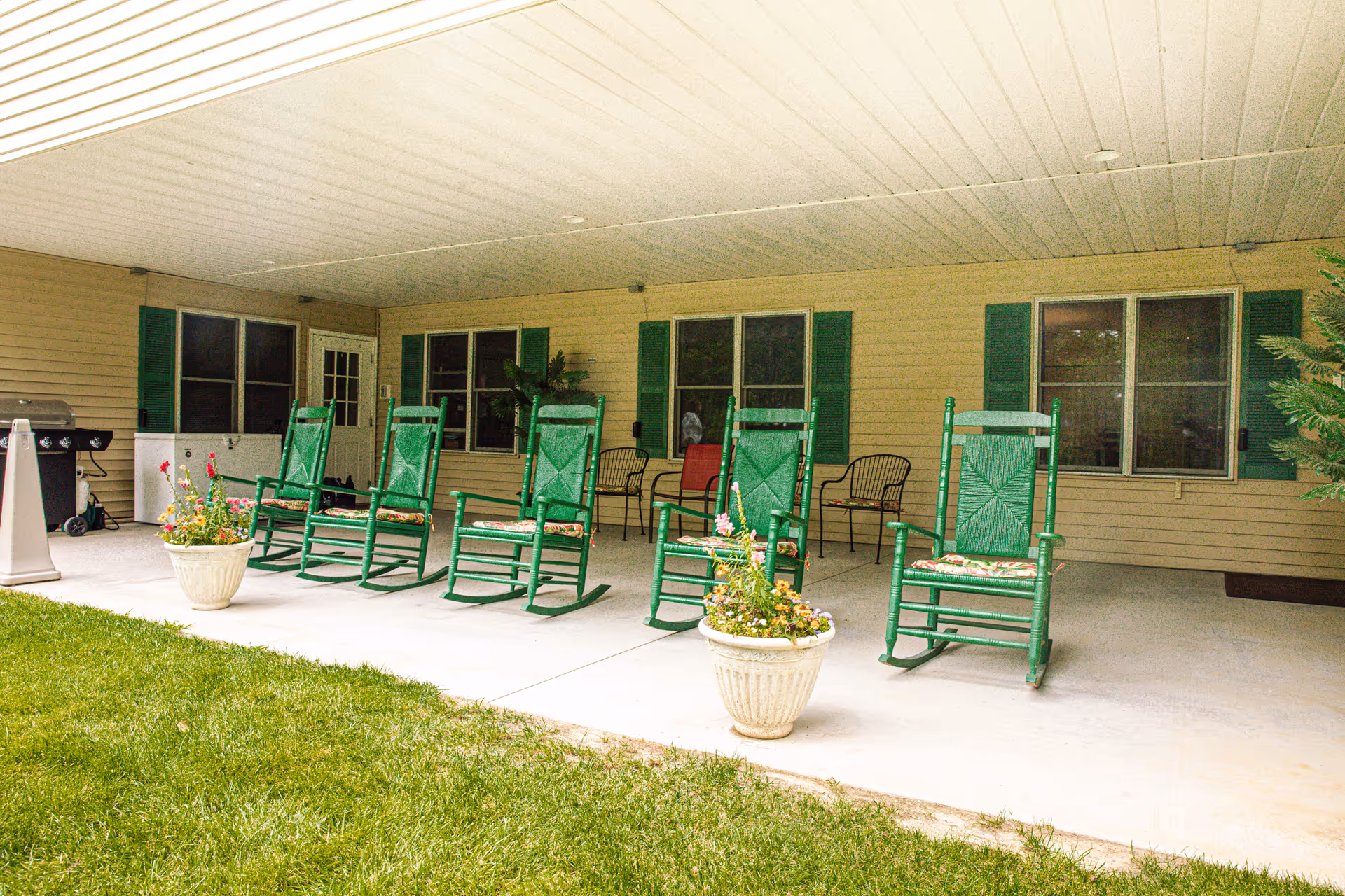 Covered outdoor patio with a row of green rocking chairs and potted flowers in front of a senior living facility.
