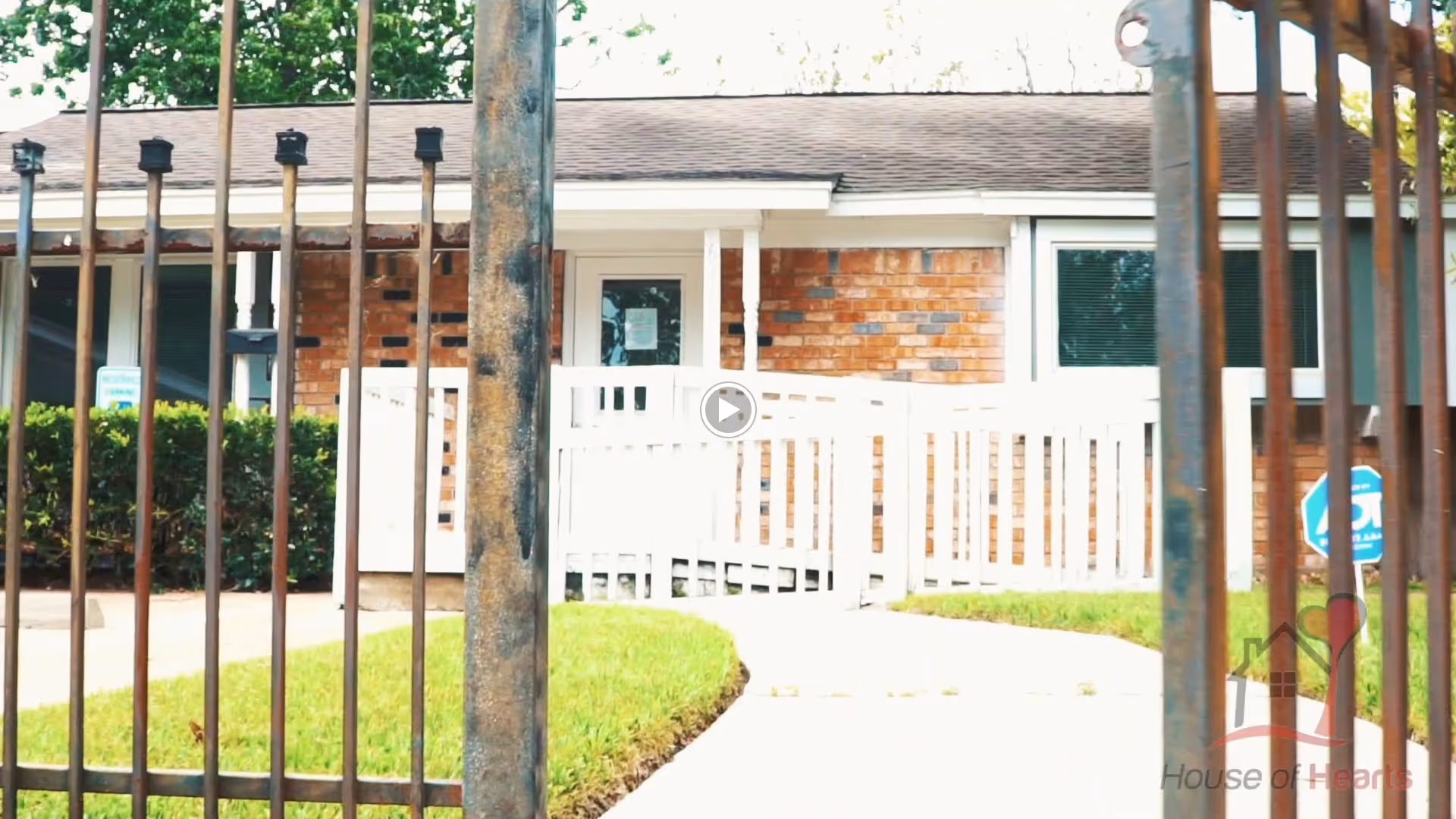 Front entrance of a single-story brick house with a white ramp and railing, seen through a metal fence.