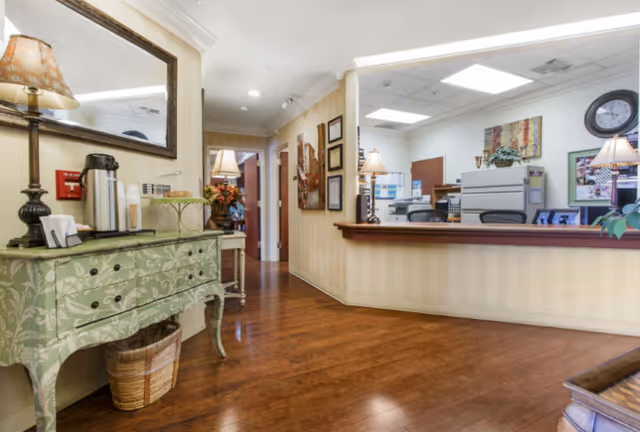 Interior view of a senior living facility reception area with a wooden floor, a green decorative console table with a lamp and coffee setup on the left, and a reception desk with office equipment and lamps in the background. The walls are light-colored with framed pictures and certificates, and there is a hallway leading further into the facility.