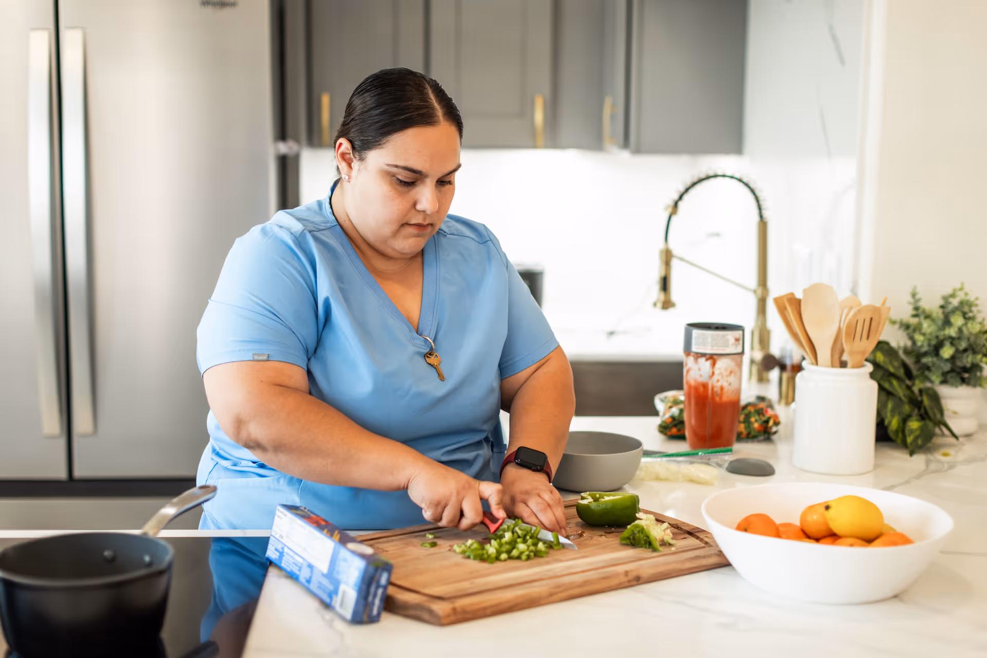 A woman in blue scrubs is chopping green bell peppers on a wooden cutting board in a modern kitchen. The kitchen features a stainless steel refrigerator, a bowl of fruit on the counter, a container with wooden utensils, and a faucet in the background.