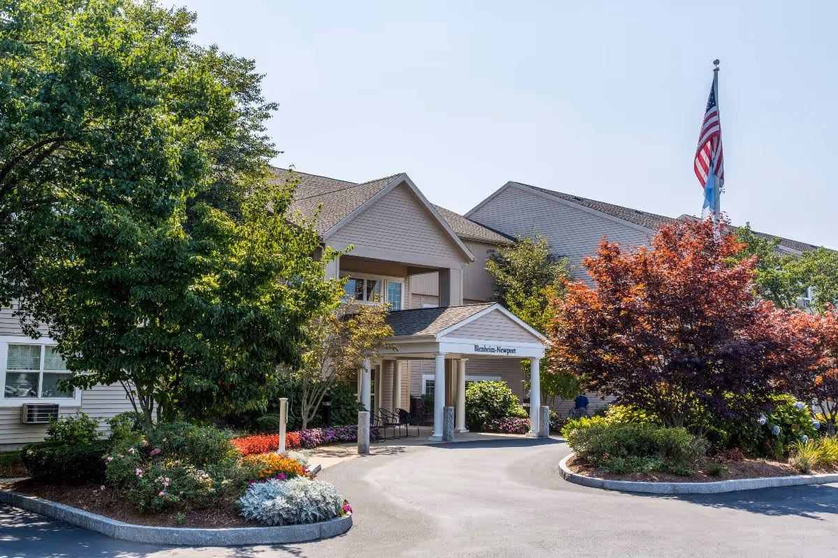 Exterior view of the Blenheim-Newport senior living facility entrance with a covered driveway, surrounded by trees, shrubs, and colorful flowers. An American flag is flying on a flagpole near the entrance.