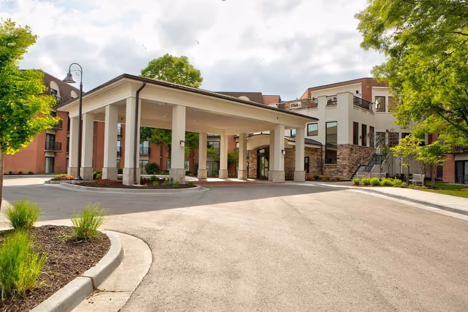 Front entrance with a covered porte-cochère and circular drive at a multi-story senior living building surrounded by trees and landscaping.