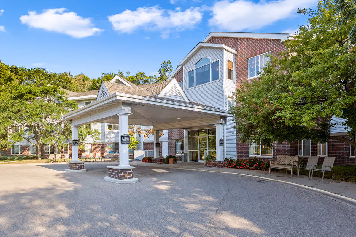 Entrance of a senior living facility named Beacon Hill with a covered drop-off area supported by white columns. The building exterior features red brick and white siding with large windows. There are trees, benches, and flowers around the entrance under a partly cloudy blue sky.