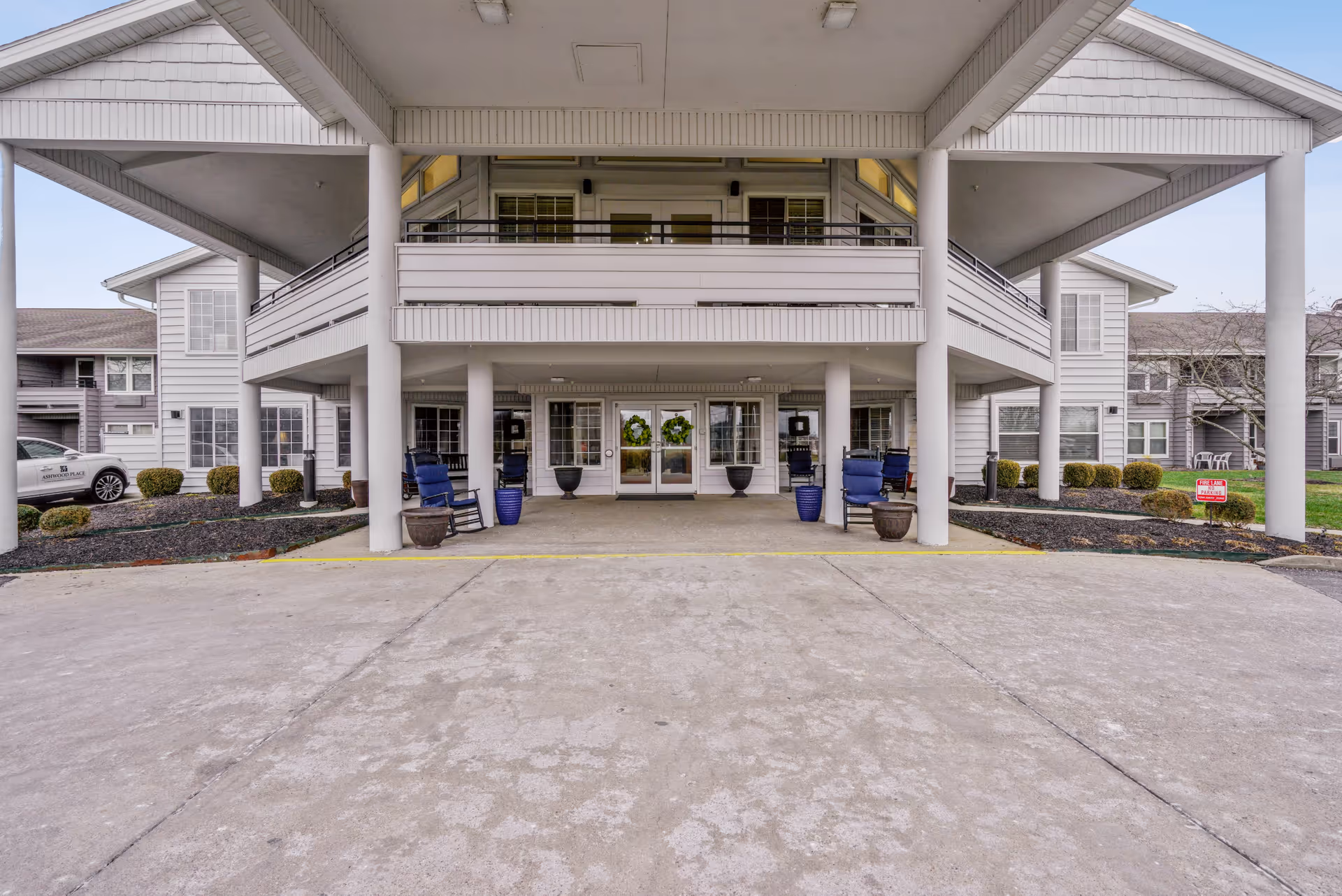 Front entrance of a senior living facility with a covered drop-off area supported by white pillars. There are blue rocking chairs and potted plants near the entrance doors. The building is two stories with balconies and multiple windows, surrounded by neatly trimmed bushes and a concrete driveway.