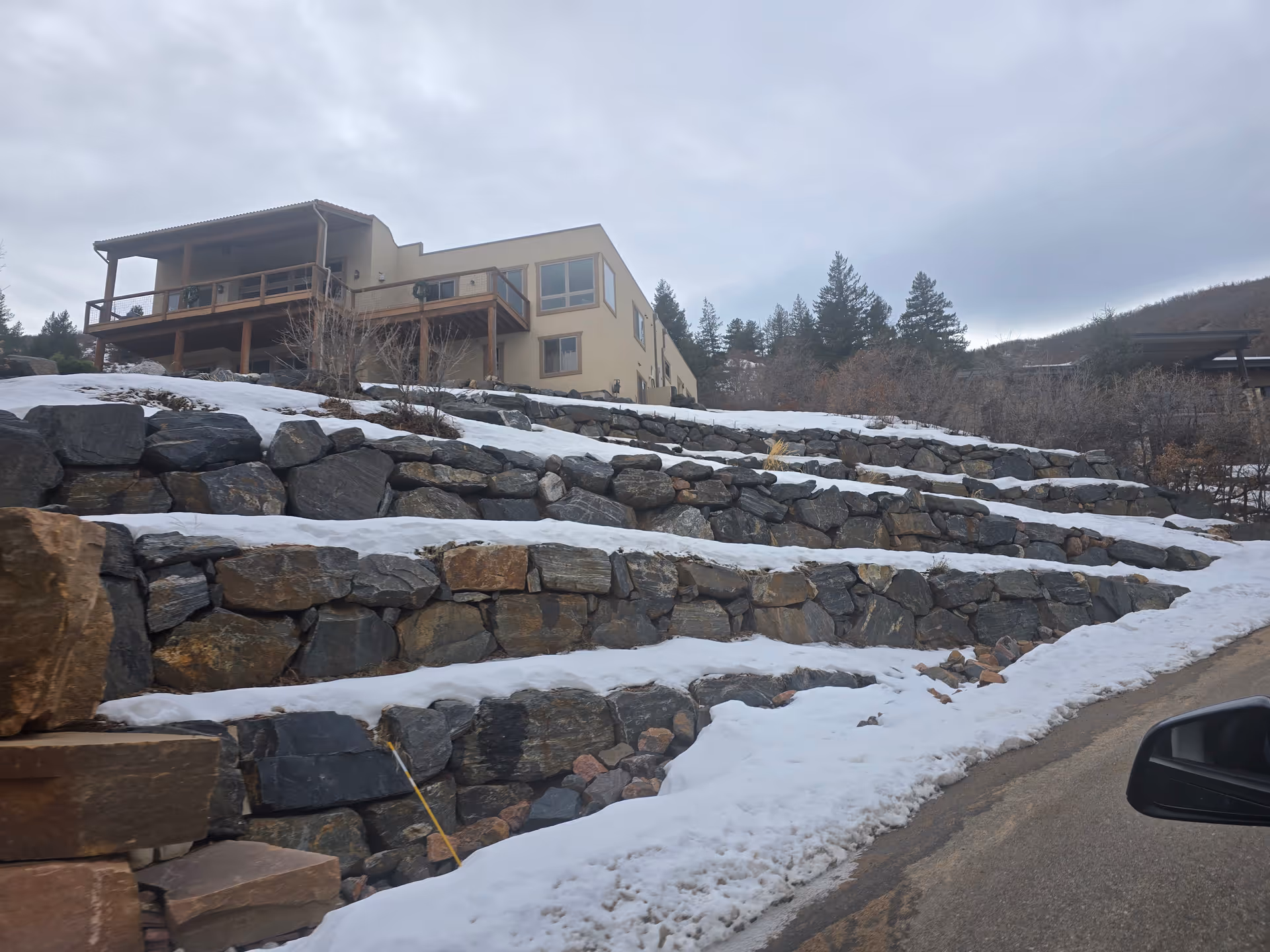 A beige building with a wooden balcony on a hillside covered with snow and large stone retaining walls. The sky is overcast and there are trees in the background. A road and a car side mirror are visible in the foreground.