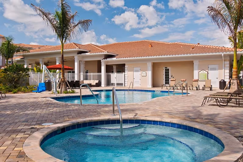 Outdoor pool area at Diamond Oaks Village with a hot tub in the foreground, a larger swimming pool behind it, lounge chairs, palm trees, and a building with a tiled roof under a partly cloudy sky.