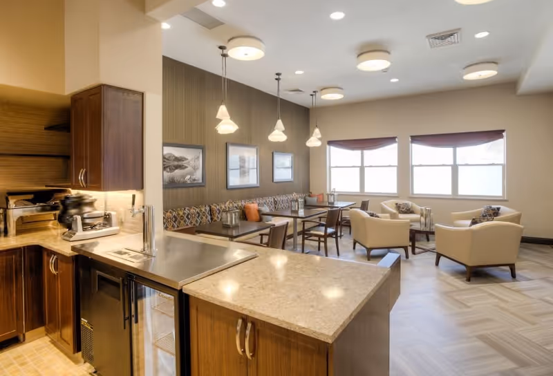 A bright and spacious common area in a senior living facility featuring a kitchen counter with a sink and mini fridge, a dining area with tables and chairs, and a lounge area with armchairs near large windows.