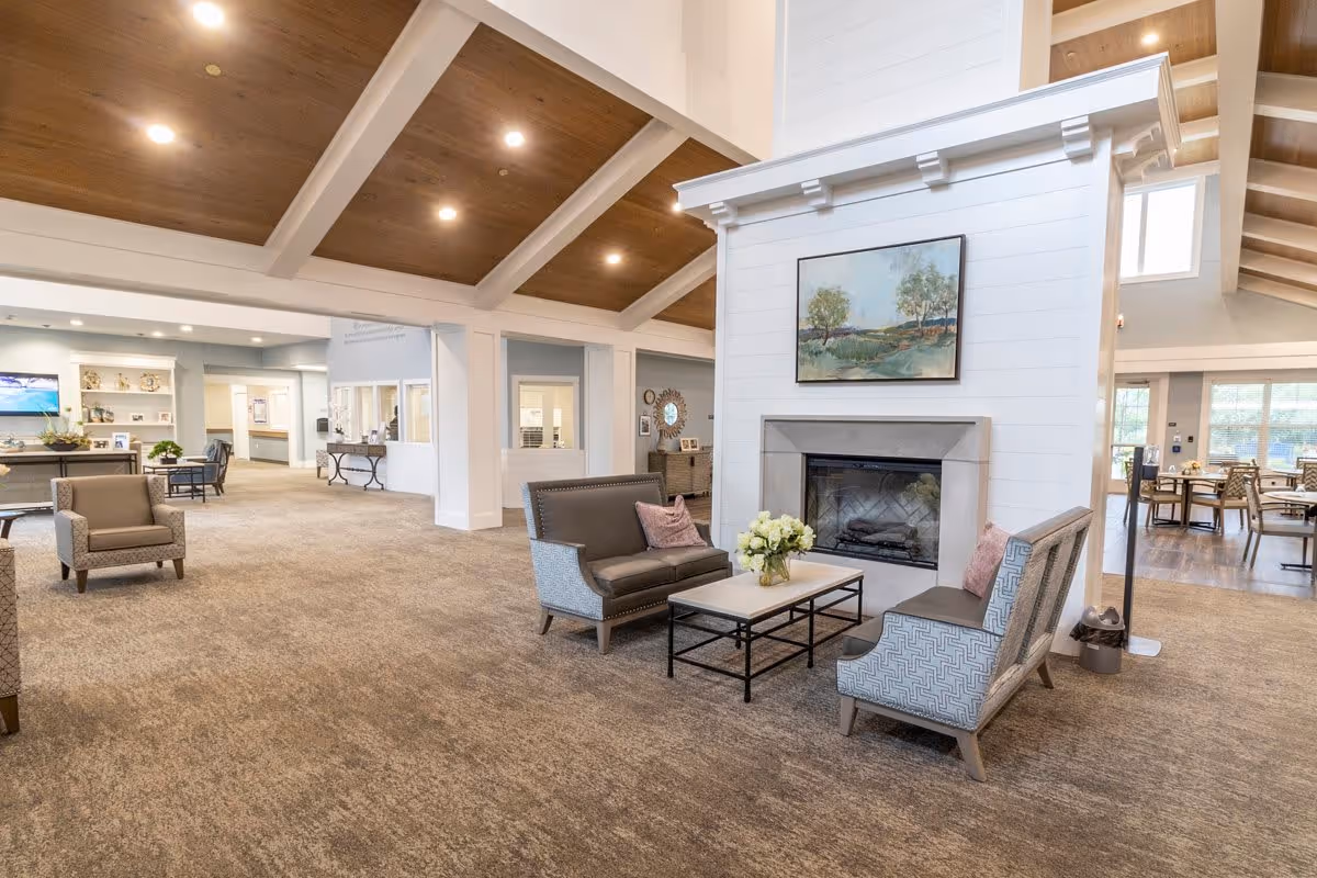 Spacious senior living facility common area with high vaulted wooden ceilings and recessed lighting. The room features a central white fireplace with a landscape painting above it, surrounded by comfortable seating including two armchairs and a loveseat with cushions, arranged around a coffee table with a flower vase. In the background, there are additional seating areas, a television, and large windows letting in natural light.