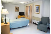 A simple, clean bedroom with a single bed covered in a light blue bedspread, a wooden nightstand with a lamp, a small wooden dresser with a flat-screen TV on top, two framed pictures on the walls, beige curtains on the window, and a gray upholstered armchair.