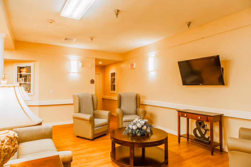 A cozy sitting area with armchairs around a coffee table topped with flowers, a wall-mounted TV, and warm lighting.