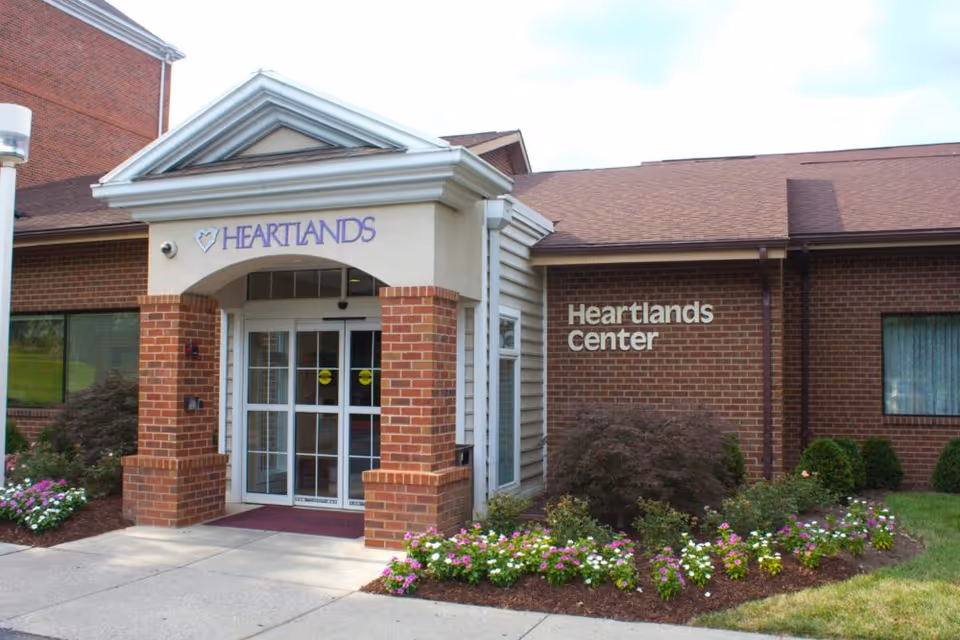 Entrance to Heartlands Center at Heart Lands Senior Living Village At Ellicott City, showing a brick building with a covered doorway, glass double doors, and landscaped flower beds on either side of the entrance.