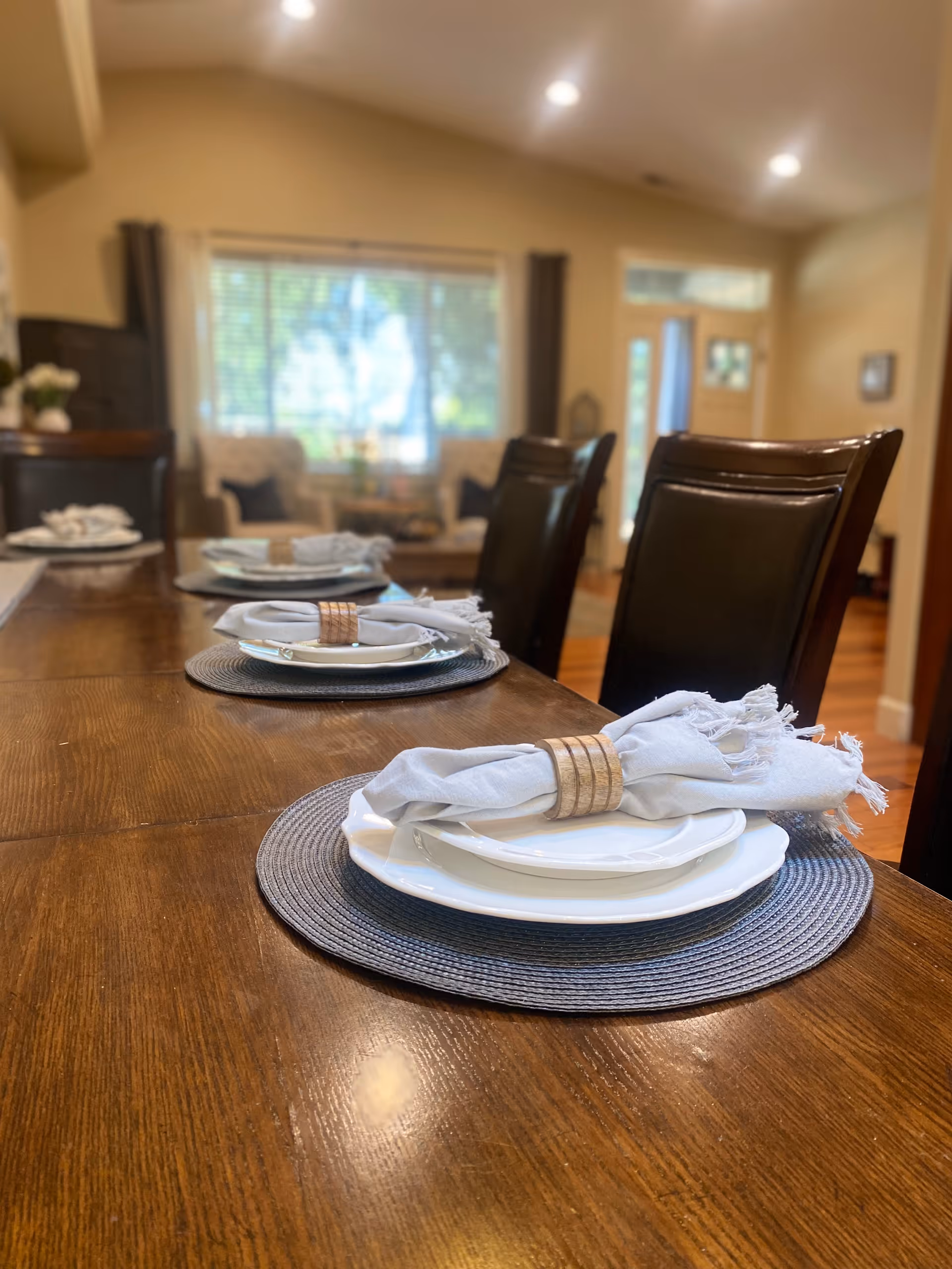A wooden dining table set with white plates, gray placemats, and light gray cloth napkins held by wooden napkin rings. The table is surrounded by dark leather chairs. In the background, there is a living area with armchairs, a window with blinds, and a door letting in natural light.