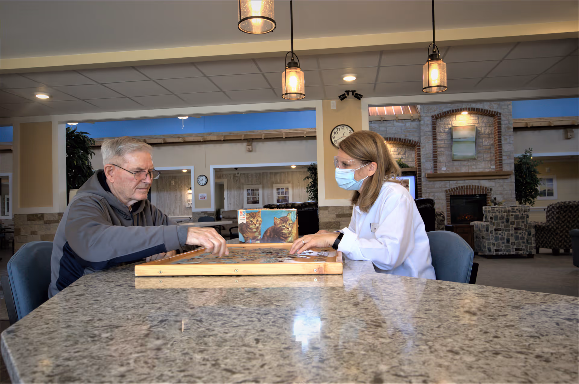 An elderly man and a masked staff member sit at a table working on a jigsaw puzzle in a communal lounge.