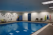 Indoor swimming pool area with clear blue water, pool safety equipment on the wall, chairs along the walls, and colorful towels hanging on hooks. The ceiling has recessed lighting and the walls are painted white.