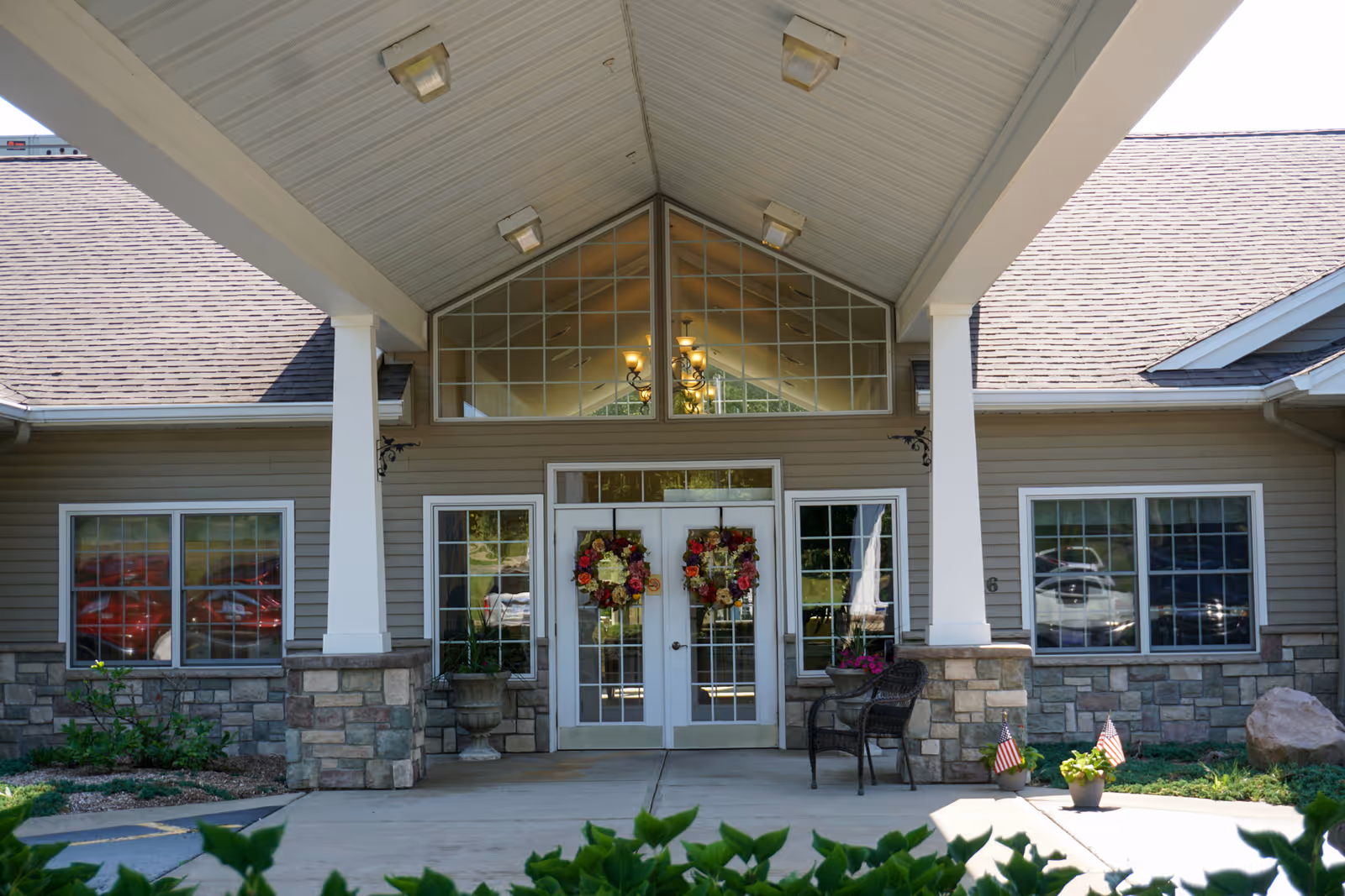 Front entrance of a senior living facility with double glass doors decorated with floral wreaths, flanked by large windows and stone pillars supporting a covered porch. There are chairs and potted plants near the entrance, and a chandelier is visible inside through the upper windows.